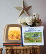Peter in Blueberry Land children's hardback book by Elsa Beskow placed on a wicker hamper next to a Toverlux lamp with coordinating artwork.
