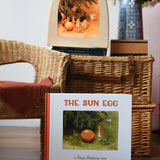 The sun egg children's hardback book by Elsa Beskow placed in front of a wicker hamper with a Toverlux lamp with sun egg art work displayed inside.