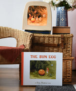 The sun egg children's hardback book by Elsa Beskow placed in front of a wicker hamper with a Toverlux lamp with sun egg art work displayed inside.