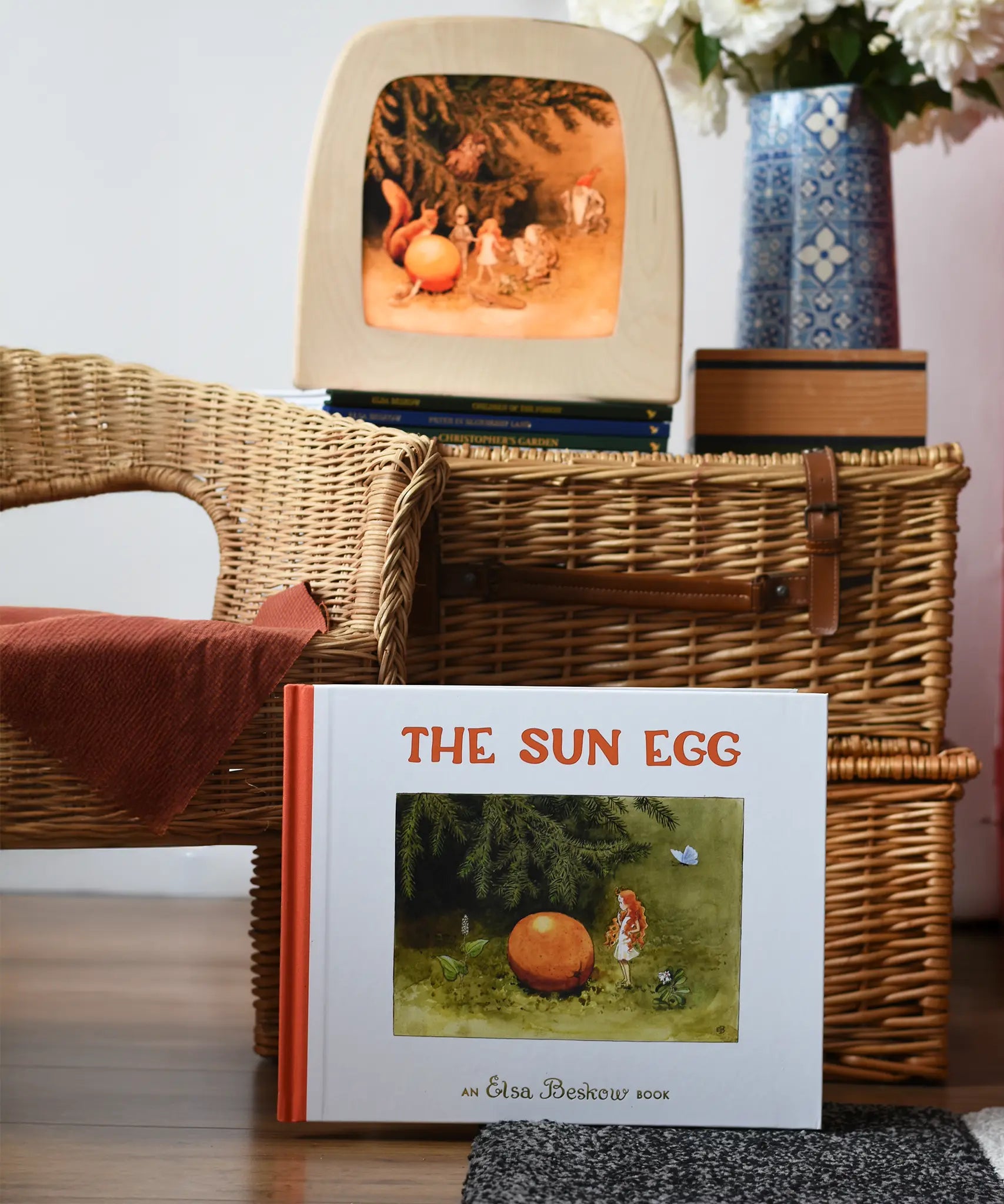 The sun egg children's hardback book by Elsa Beskow placed in front of a wicker hamper with a Toverlux lamp with sun egg art work displayed inside.