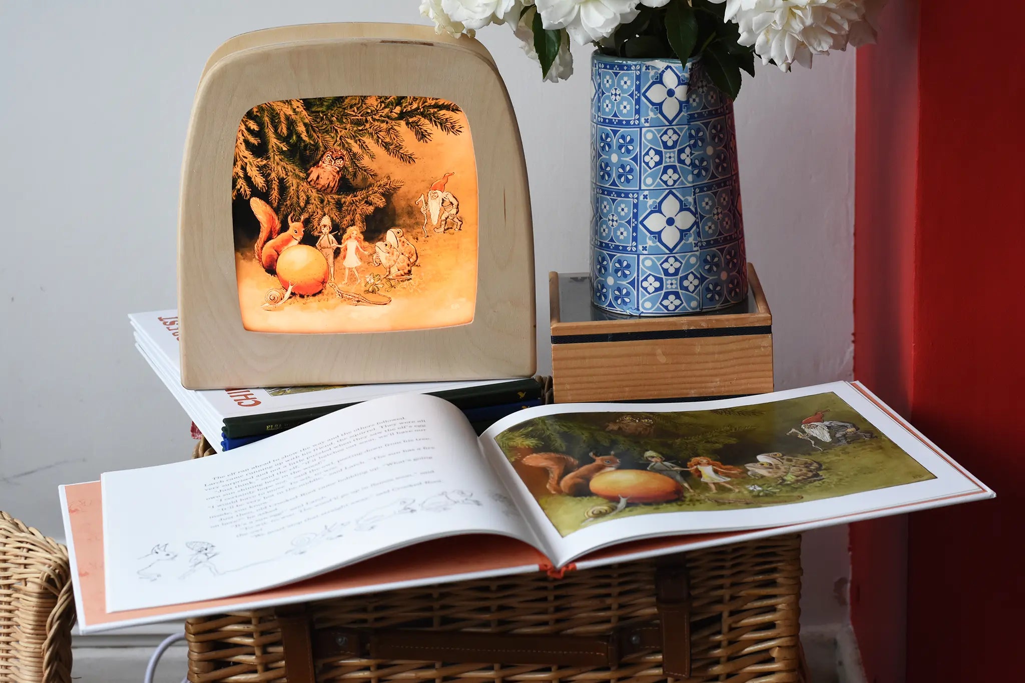 The sun egg children's hardback book by Elsa Beskow shown open placed on a wicker hamper with a Toverlux lamp with sun egg art work displayed inside.