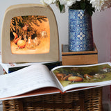 The sun egg children's hardback book by Elsa Beskow shown open placed on a wicker hamper with a Toverlux lamp with sun egg art work displayed inside.