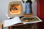 The sun egg children's hardback book by Elsa Beskow shown open placed on a wicker hamper with a Toverlux lamp with sun egg art work displayed inside.