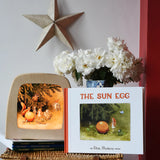 The sun egg children's hardback book by Elsa Beskow placed on a wicker hamper with a Toverlux lamp with sun egg art work displayed inside.