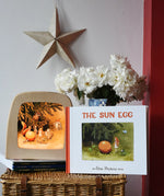 The sun egg children's hardback book by Elsa Beskow placed on a wicker hamper with a Toverlux lamp with sun egg art work displayed inside.