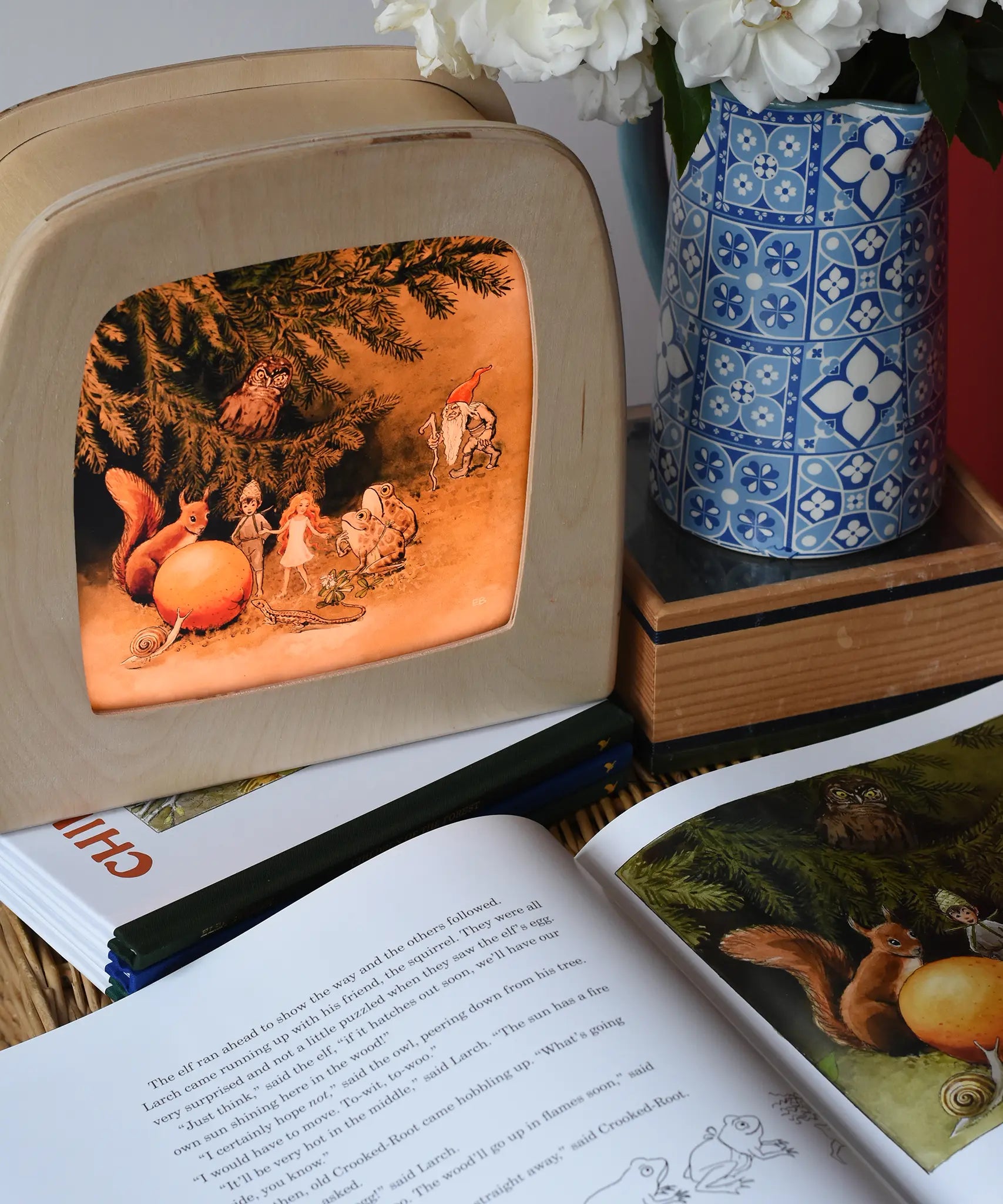 The sun egg children's hardback book by Elsa Beskow shown open placed on a wicker hamper with a Toverlux lamp with sun egg art work displayed inside.