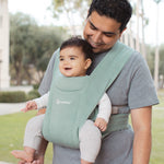 Man wearing a grey t-shirt and his baby on his front facing outwards in the Ergobaby Embrace Soft Knit Newborn Baby Carrier in Jade. Side view in a garden.
