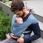 Man wearing a navy blue top and his baby on his front facing inwards in the Ergobaby Embrace Soft Knit Newborn Baby Carrier in Oxford Blue. Side view sat on a garden bench.