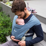 Man wearing a navy blue top and his baby on his front facing inwards in the Ergobaby Embrace Soft Knit Newborn Baby Carrier in Oxford Blue. Side view sat on a garden bench.