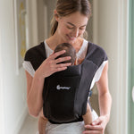 Woman wearing a white t-shirt and her baby on her front in the Ergobaby Embrace Soft Knit Newborn Baby Carrier in Pure Black. Front view in a hallway.
