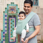Man wearing a grey t-shirt and his baby on his front facing outwards in the Ergobaby Embrace Soft Knit Newborn Baby Carrier in Jade. Side view in front of a colourful tiled wall.