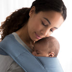 Woman wearing a grey sweater and her sleeping baby on her front in the Ergobaby Embrace Soft Knit Newborn Baby Carrier in Oxford Blue. Side view white background.