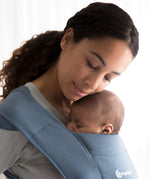 Woman wearing a grey sweater and her sleeping baby on her front in the Ergobaby Embrace Soft Knit Newborn Baby Carrier in Oxford Blue. Side view white background.