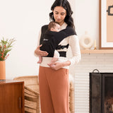 Woman wearing a white top and brown trousers and her baby on her front in the Ergobaby Embrace Soft Knit Newborn Baby Carrier in Pure Black. Front view in front of a white tiled fireplace.