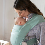 Woman wearing a floral blouse and her baby on her front facing inwards in the Ergobaby Embrace Soft Knit Newborn Baby Carrier in Jade. Side view next to a cot.