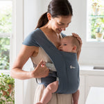 Woman wearing a white vest and her baby on her front facing inwards in the Ergobaby Embrace Soft Knit Newborn Baby Carrier in Oxford Blue. Side view, looking down to stroke baby's head.