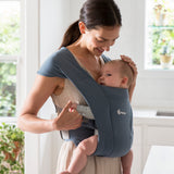 Woman wearing a white vest and her baby on her front facing inwards in the Ergobaby Embrace Soft Knit Newborn Baby Carrier in Oxford Blue. Side view, looking down to stroke baby's head.