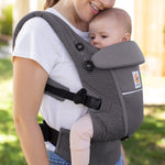 Woman with brown hair and a white t-shirt wearing her baby in the Ergobaby Adapt  SoftFlex™ Mesh Baby Carrier in Graphite Grey. Front view, outside in a garden. 