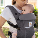 Woman with brown hair and a white t-shirt wearing her baby in the Ergobaby Adapt  SoftFlex™ Mesh Baby Carrier in Graphite Grey. Front view, outside in a garden. 