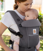 Woman with brown hair and a white t-shirt wearing her baby in the Ergobaby Adapt  SoftFlex™ Mesh Baby Carrier in Graphite Grey. Front view, outside in a garden. 