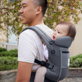 Man with black hair and a white t-shirt wearing his baby on his back in the Ergobaby Adapt  SoftFlex™ Mesh Baby Carrier in Graphite Grey. Side view, outside in a garden. 