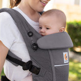 Woman with brown hair and a white t-shirt wearing her baby in the Ergobaby Adapt  SoftFlex™ Mesh Baby Carrier in Graphite Grey. Front view, outside in a garden. 