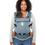 Woman with black curly hair and a grey jumper wearing her baby in the Ergobaby Adapt  SoftFlex™ Mesh Baby Carrier in Slate Blue. Front view, white background. 