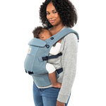 Woman with black curly hair and a grey jumper wearing her baby in the Ergobaby Adapt  SoftFlex™ Mesh Baby Carrier in Slate Blue. Side view, white background. 