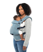 Woman with black curly hair and a grey jumper wearing her baby in the Ergobaby Adapt  SoftFlex™ Mesh Baby Carrier in Slate Blue. Side view, white background. 