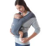 Woman wearing a grey top and her baby on her front facing inwards in the Ergobaby Embrace Soft Knit Newborn Baby Carrier in Oxford Blue. Side view, white background.