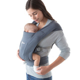 Woman wearing a grey top and her baby on her front facing inwards in the Ergobaby Embrace Soft Knit Newborn Baby Carrier in Oxford Blue. Side view, white background.