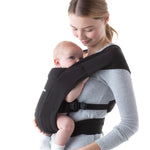 Woman wearing a grey top and her baby on her front in the Ergobaby Embrace Soft Knit Newborn Baby Carrier in Pure Black. Side view white background.