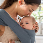 Woman wearing a white vest and her baby on her front facing inwards in the Ergobaby Embrace Soft Knit Newborn Baby Carrier in Oxford Blue. Side view, leaning forward to kiss baby's head.