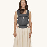 A woman wearing the Ergobaby Omni Deluxe Mesh baby carrier in Graphite Grey, with a little baby being carried facing in towards her chest. 