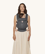 A woman wearing the Ergobaby Omni Deluxe Mesh baby carrier in Graphite Grey, with a little baby being carried facing in towards her chest. 