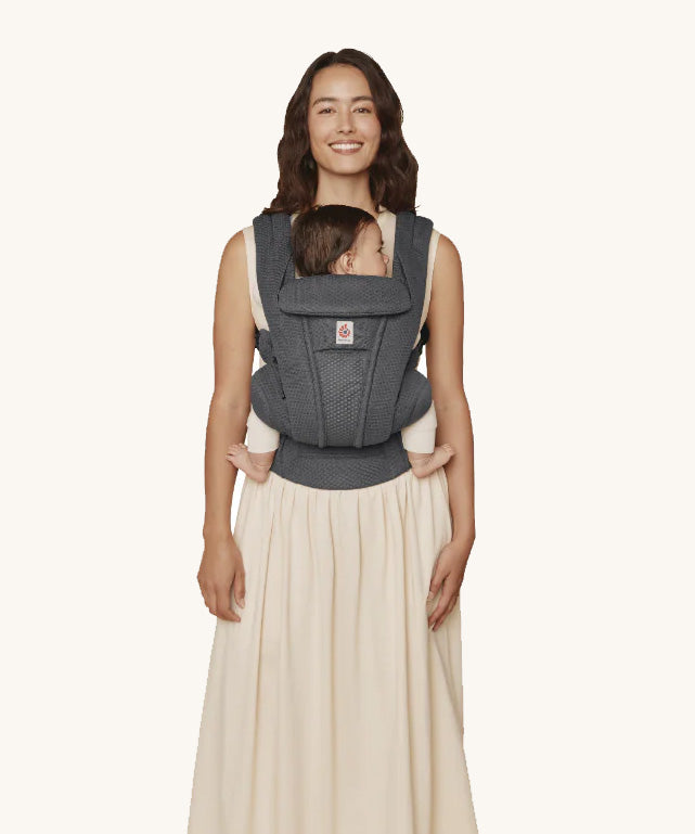 A woman wearing the Ergobaby Omni Deluxe Mesh baby carrier in Graphite Grey, with a little baby being carried facing in towards her chest. 