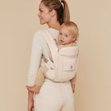 A woman wearing a baby on her back with the Ergobaby Omni Deluxe Cotton baby carrier in Natural Beige.