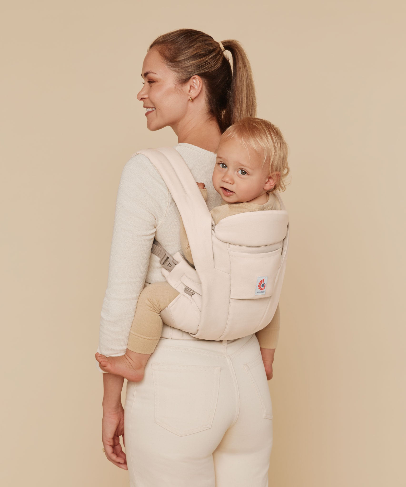 A woman wearing a baby on her back with the Ergobaby Omni Deluxe Cotton baby carrier in Natural Beige.