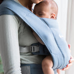 Woman wearing a grey top and her sleeping baby on her front facing inwards in the Ergobaby Embrace Soft Knit Newborn Baby Carrier in Oxford Blue. Side view in front of a window.