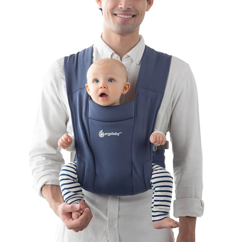A man carrying a small baby in the Ergobaby soft knit newborn baby carrier, in soft navy.