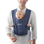 A man carrying a small baby in the Ergobaby soft knit newborn baby carrier, in soft navy.