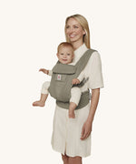 A woman wearing the Ergobaby Omni Deluxe Mesh baby carrier in Soft Olive, with a little baby being carried facing outwards. 