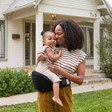 A woman carrying her little girl on the Ergobaby Lift Hip Seat baby carrier, outside a house.