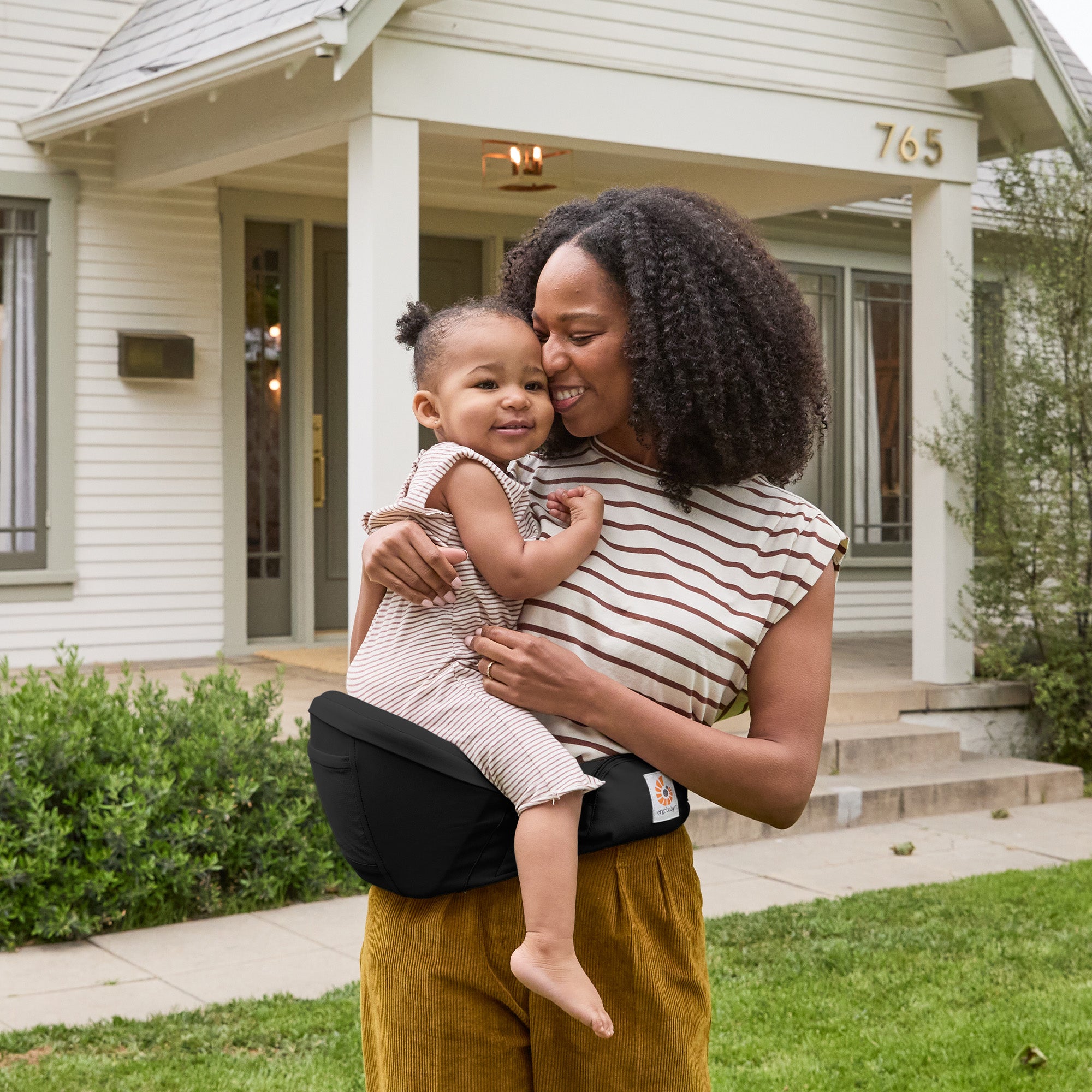 A woman carrying her little girl on the Ergobaby Lift Hip Seat baby carrier, outside a house.