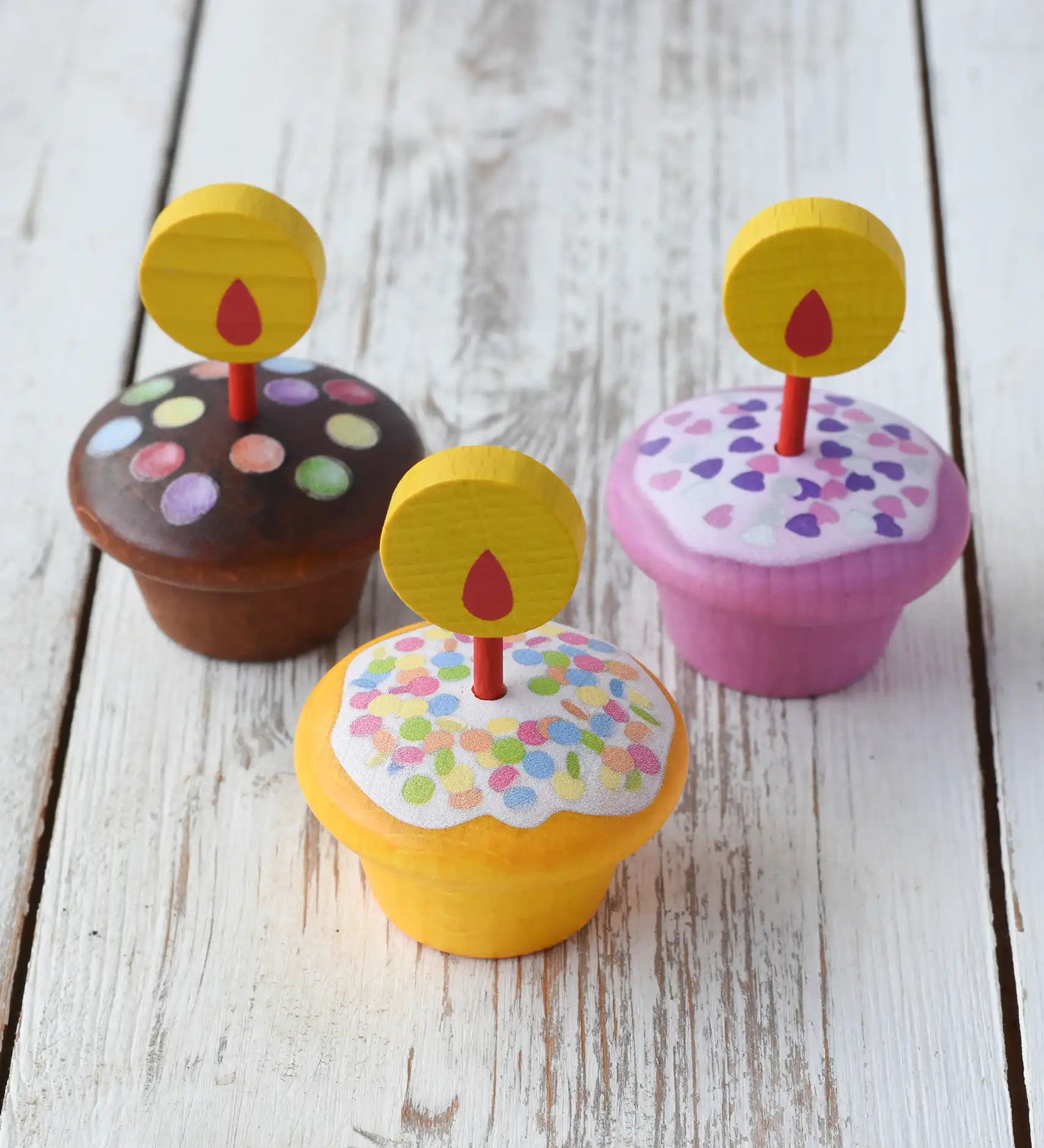 Erzi Birthday Muffins Wooden Play Food. Set of three small toy cupcakes with colorful sprinkles and yellow flame candle toppers, arranged on a whitewashed wooden surface.