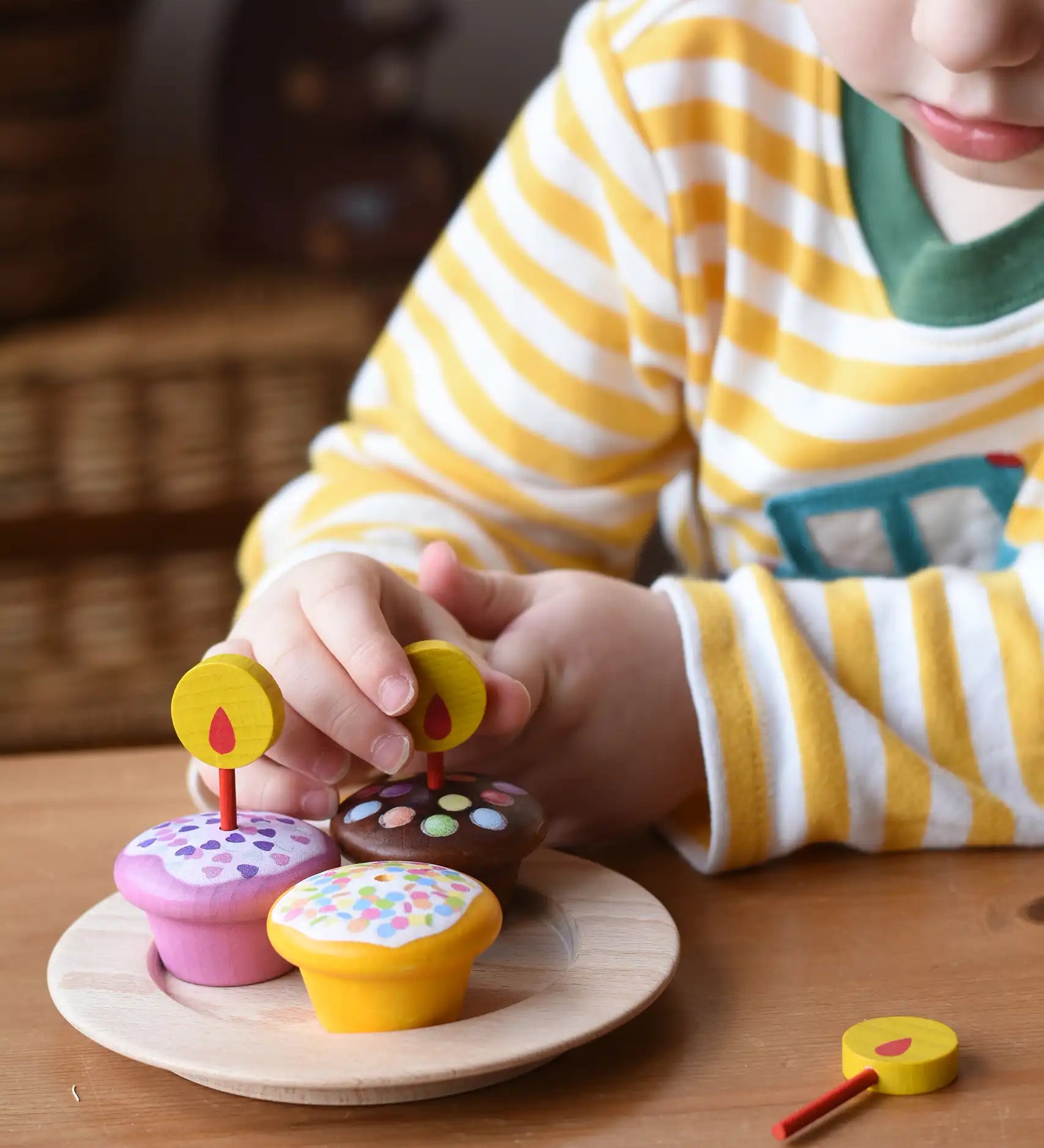 Erzi Birthday Muffins Wooden Play Food. Set of three small toy cupcakes with colorful sprinkles and yellow flame candle toppers, arranged on a wooden plate with a child placing a candle on one of the cakes.