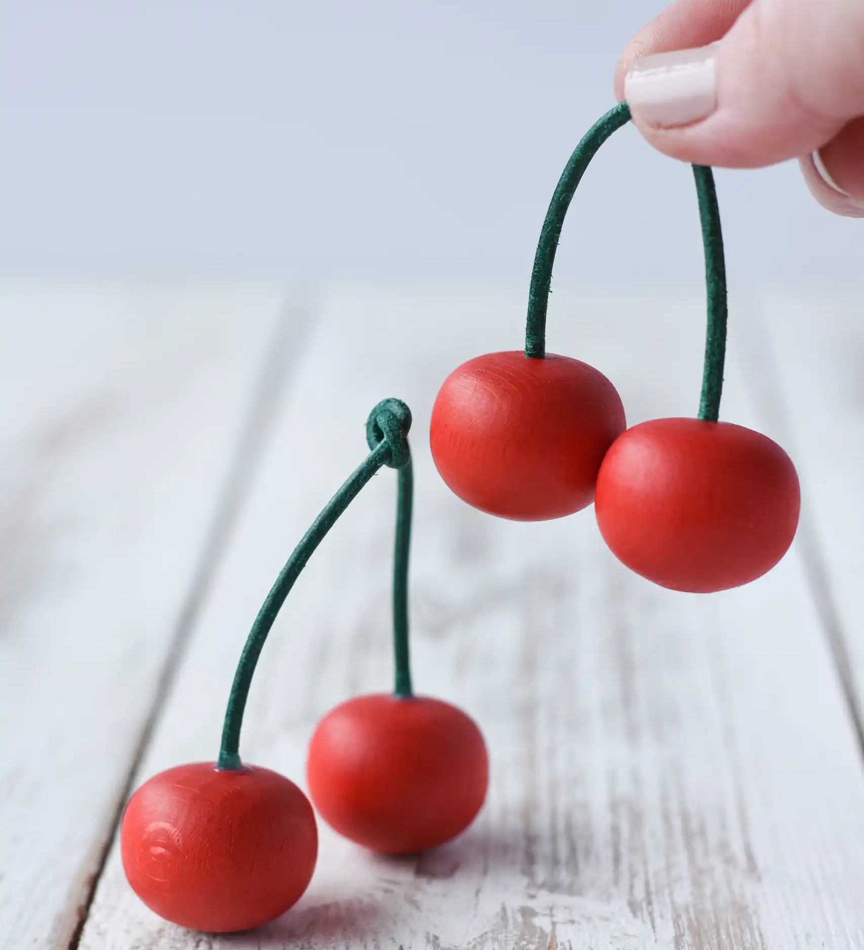 Erzi pair of cherries wooden play food placed on a wooden surface with another pair in an adult's hand. Red fruits on a green stem.