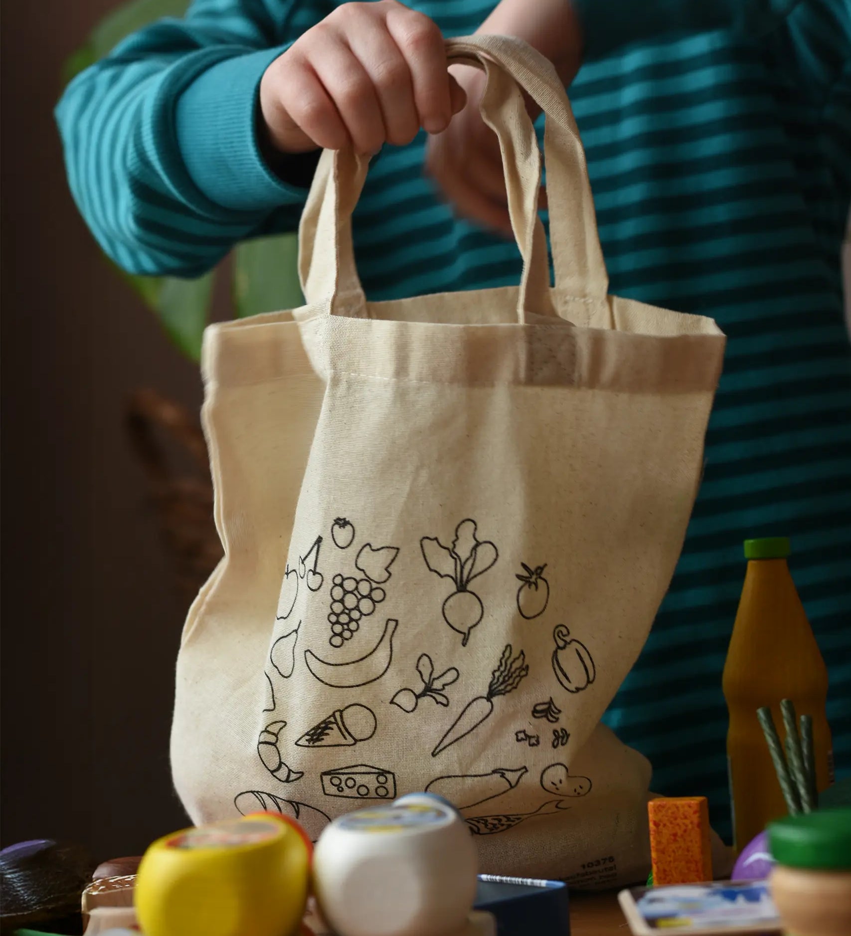Erzi cotton shopping tote bag in a child's hand. The child is emptying the toy play food from inside.