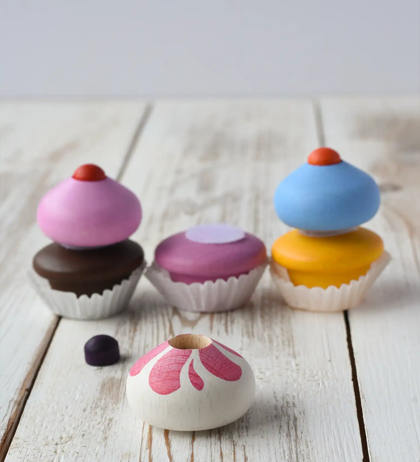 Three Erzi colorful wooden toy cupcakes with removable frosting tops, sprinkle and paper cup liners, arranged on a white rustic wooden surface. One cupcake has the frosting and sprinkle removed.