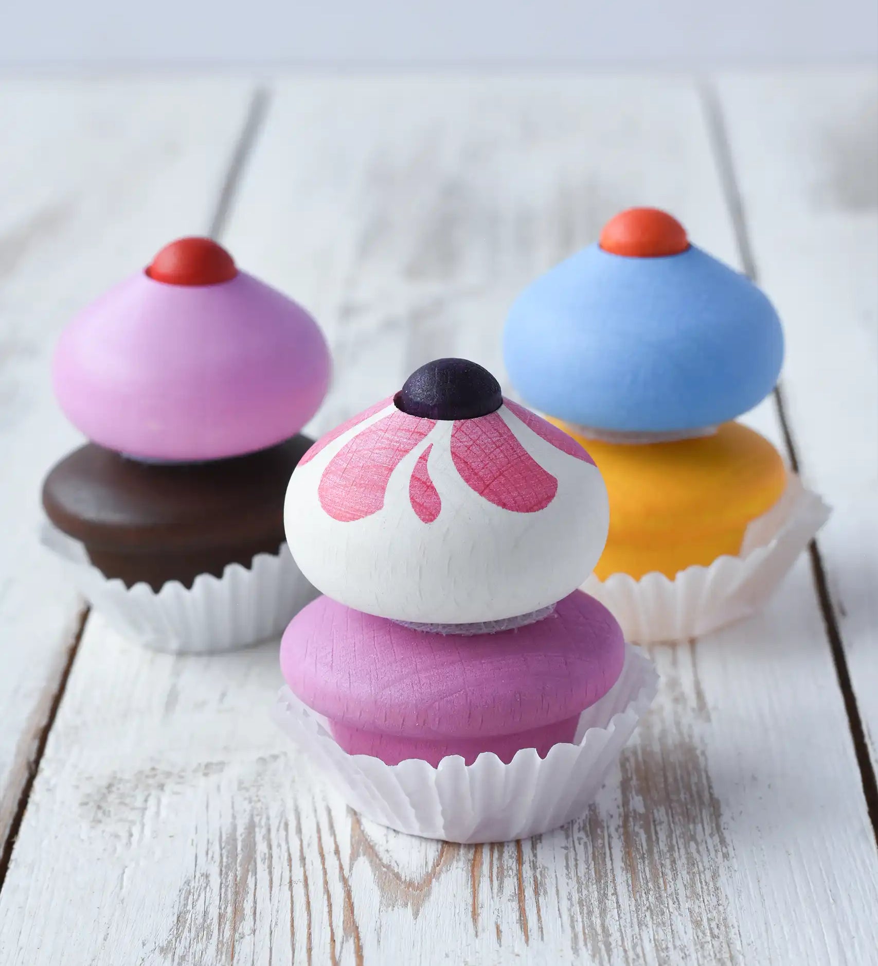 Three Erzi colorful wooden toy cupcakes with removable frosting tops, sprinkle and paper cup liners, arranged on a white rustic wooden surface.
